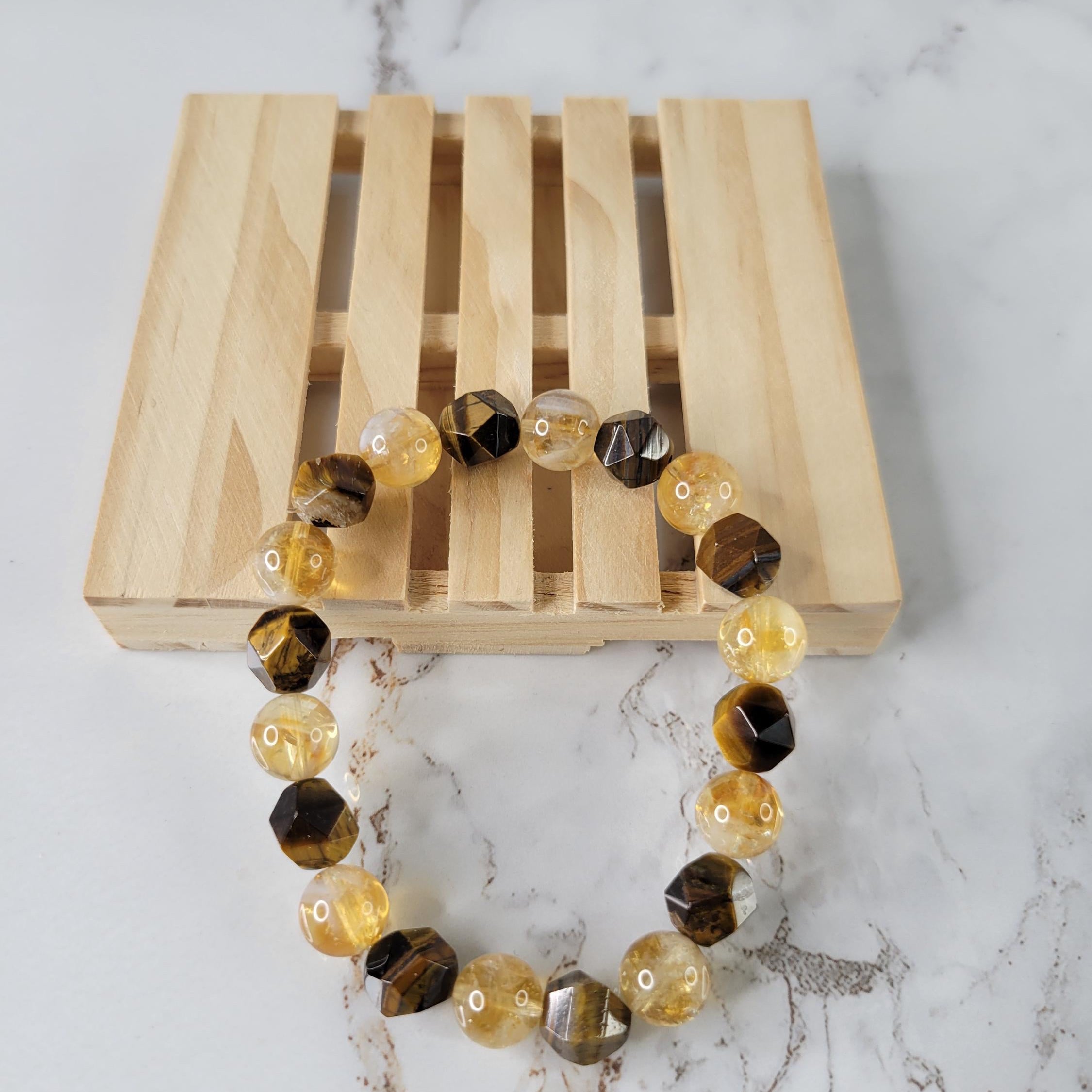 A beaded stretch bracelet made of faceted translucent yellow beads and dark brown tiger's eye beads, displayed on a light wood slatted stand over a white marble surface.
