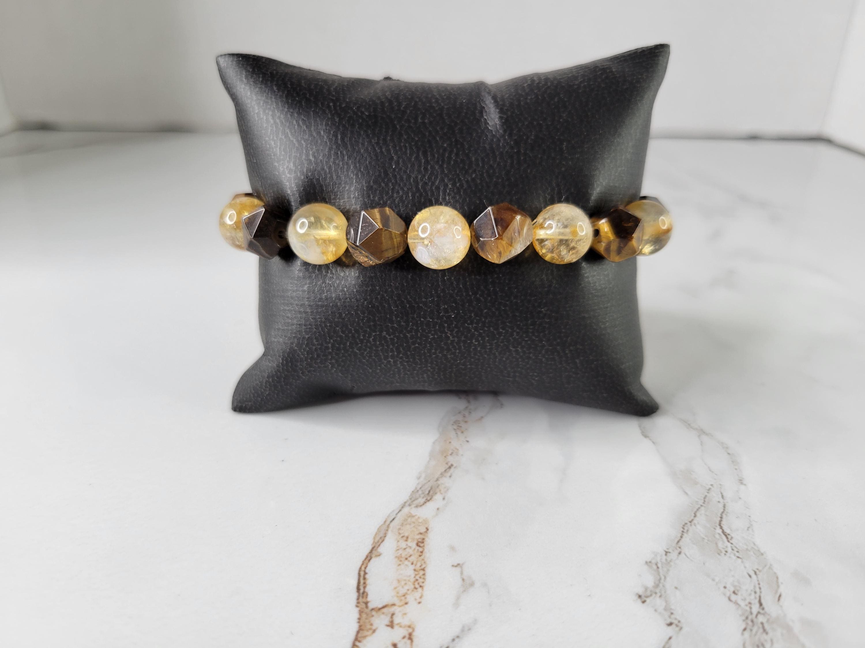 A beaded stretch bracelet with faceted amber, gold, and iridescent beads, displayed on a black faux-leather pillow over a white marble surface.