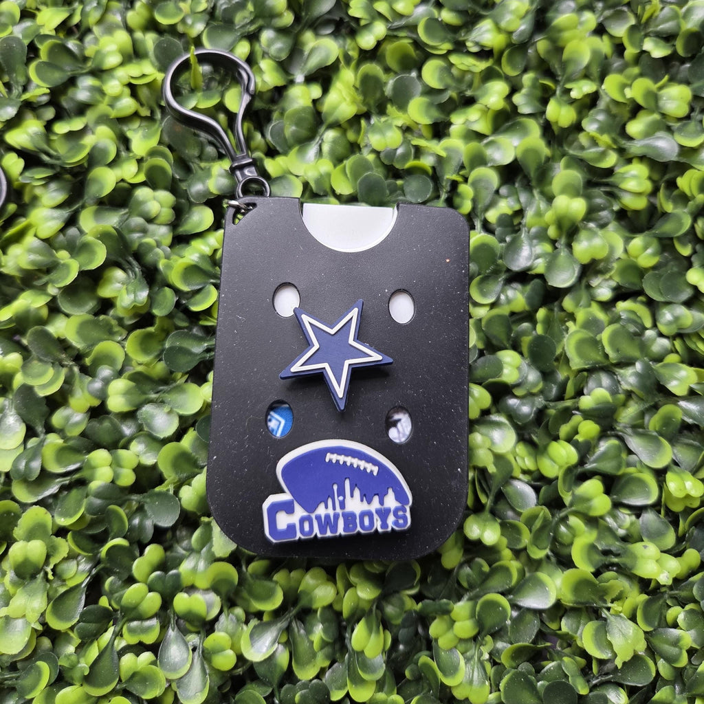 Dallas Cowboys themed keychain featuring a black rectangular card with a blue star and Cowboys logo, attached to a black key ring, displayed on a green leafy background.
