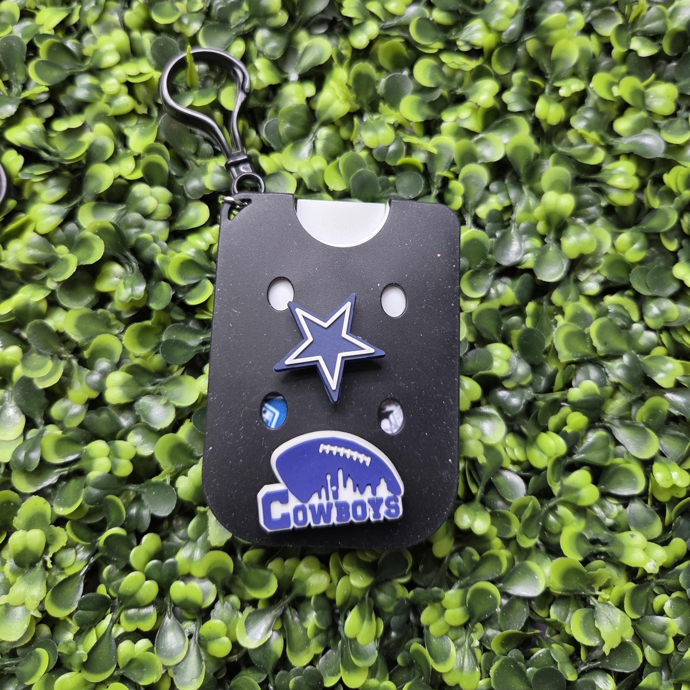 Dallas Cowboys themed keychain featuring a black rectangular card with a blue star and Cowboys logo, attached to a black key ring, displayed on a green leafy background.