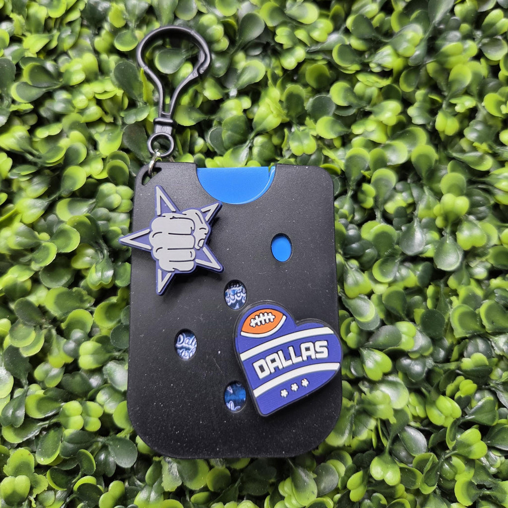 A black rectangular keychain or card holder with a metal clip, decorated with a silver-and-blue star-shaped charm, a blue circular piece, and a Dallas Cowboys football-themed patch on a green leafy background.