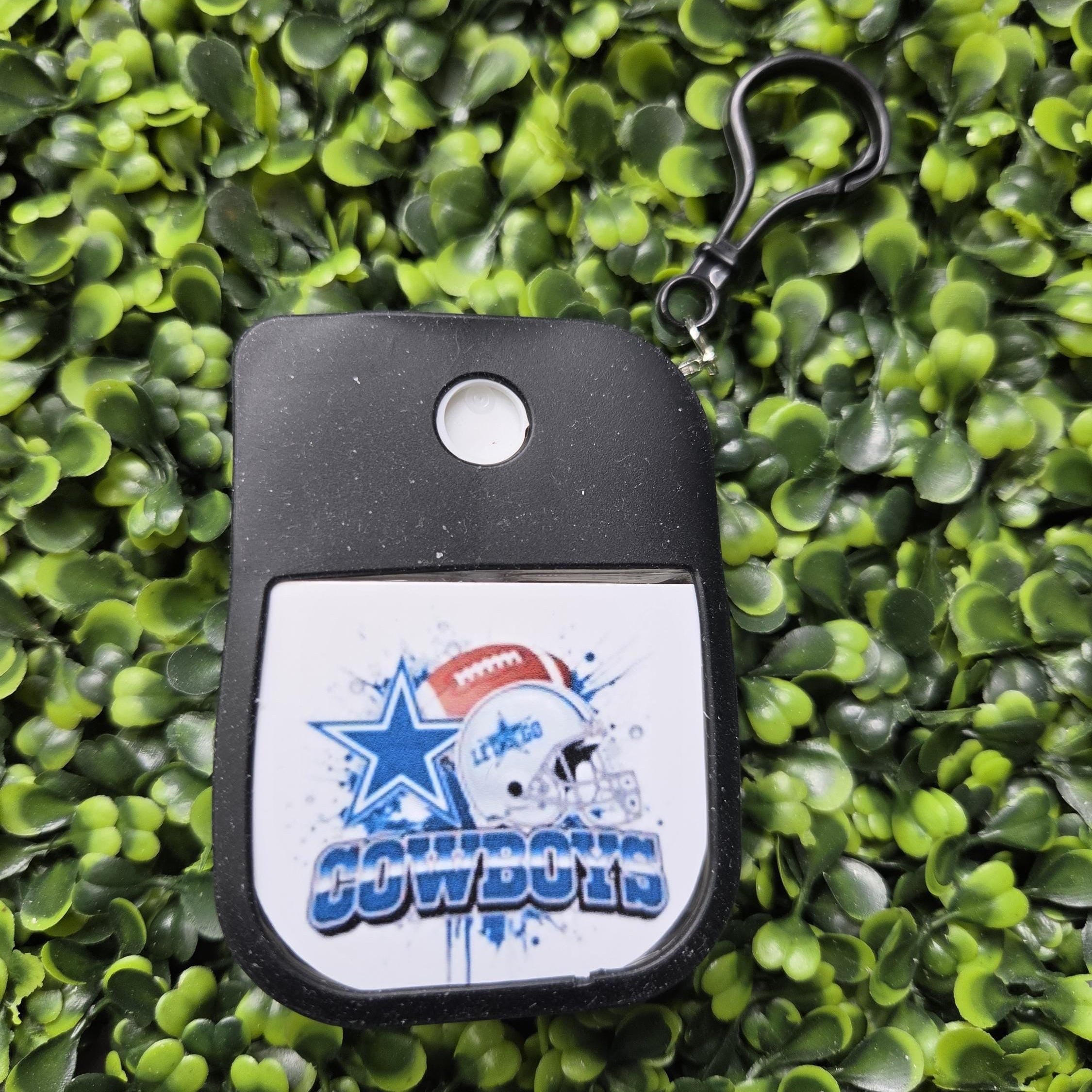A Dallas Cowboys themed key fob or alarm key tag with a black plastic body and a white graphic insert featuring the team logo and name, attached to a black carabiner clip, set against a background of green artificial foliage.