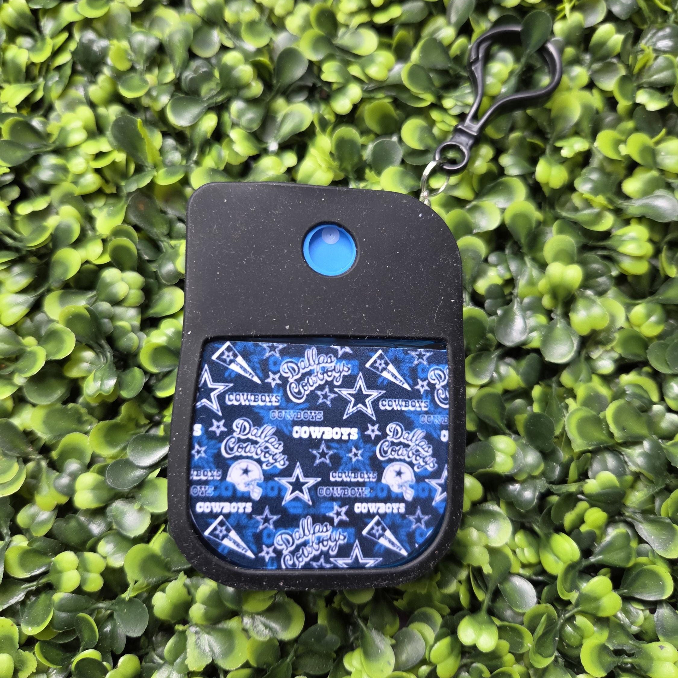 A Dallas Cowboys themed card holder or pocket clip with a black silicone body, a blue circular button, and a blue patterned front featuring the Cowboys logo, placed on a green leafy background.
