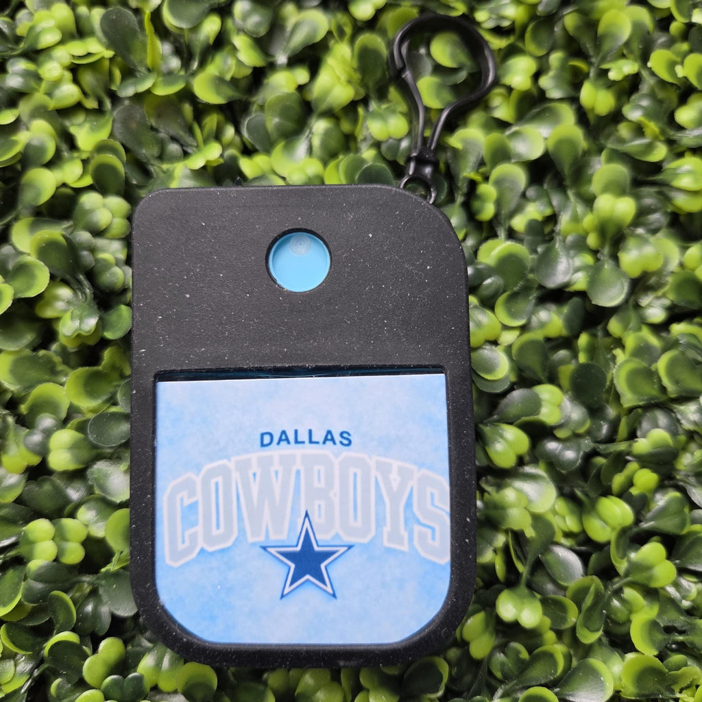 A Dallas Cowboys themed keychain or alarm tag with a black body, a light blue circular button, and a light blue front panel featuring the Dallas Cowboys logo and text, shown against a background of artificial green foliage.