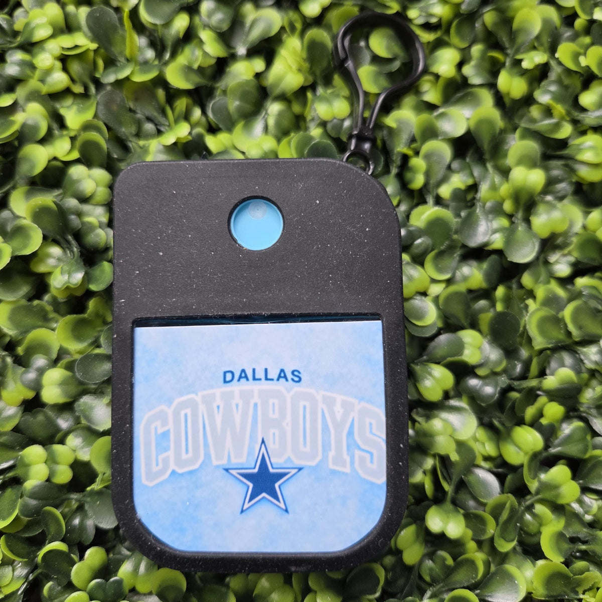 A Dallas Cowboys themed keychain or alarm tag with a black body, a light blue circular button, and a light blue front panel featuring the Dallas Cowboys logo and text, shown against a background of artificial green foliage.