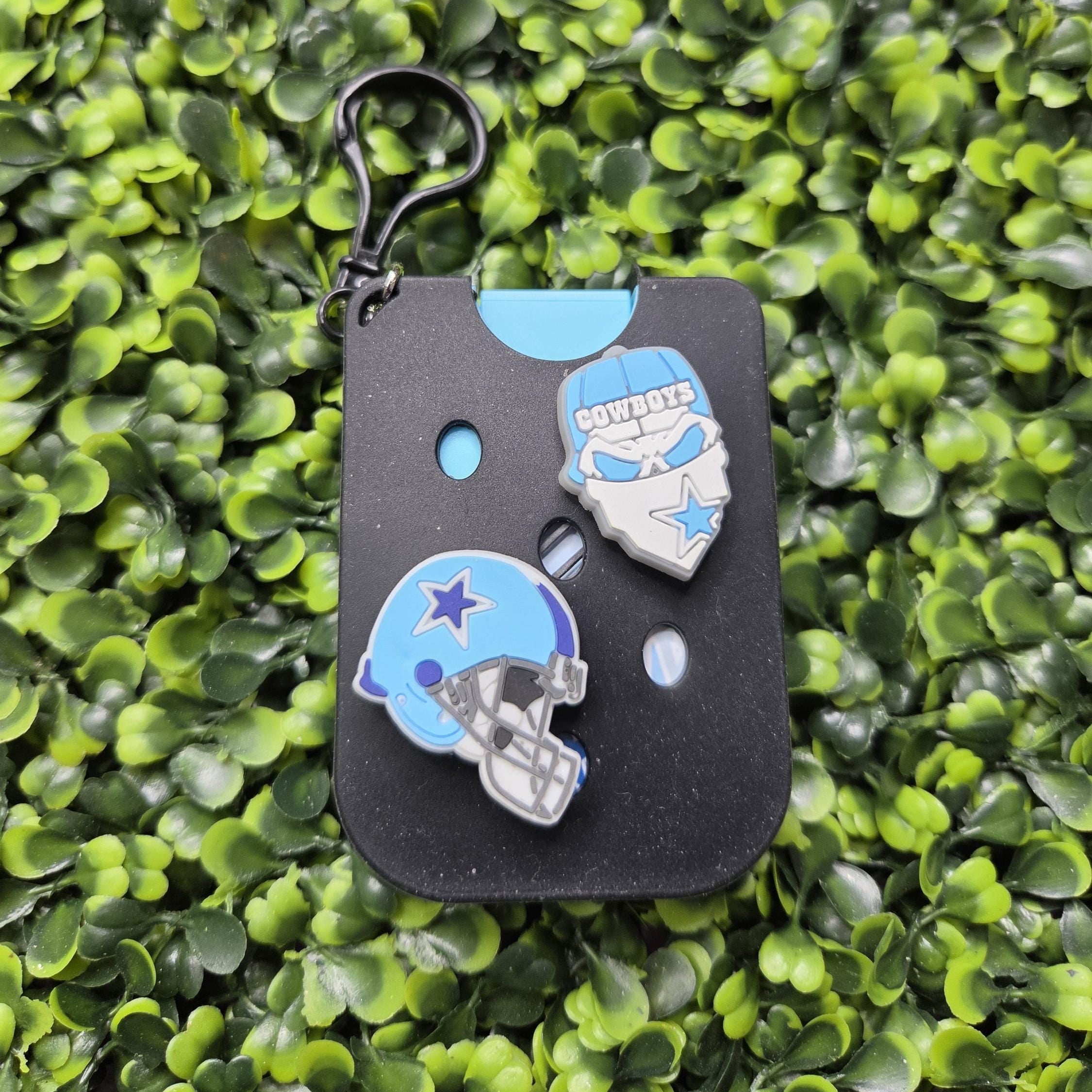 A Dallas Cowboys themed keychain or bag charm set displayed on a black backing card, featuring a football helmet and a star logo, set against a background of artificial green foliage.